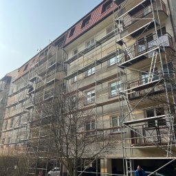 TDL GROUP SP.J. - Wielopiętrowy budynek mieszkalny w trakcie prac elewacyjnych, osłonięty rusztowaniami. Widoczny pracownik w odblaskowej odzieży. Słoneczny dzień.