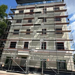 TDL GROUP SP.J. - Wielopiętrowy budynek w trakcie prac elewacyjnych, osłonięte okna, rusztowania na całej wysokości, widok z dołu, błękitne niebo z chmurami w tle.