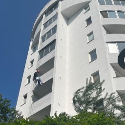 TDL GROUP SP.J. - Pracownik na linach, w kasku, maluje białą elewację wysokiego budynku z balkonami. Widok z dołu, błękitne niebo w tle, zieleń drzew w dolnej części kadru.
