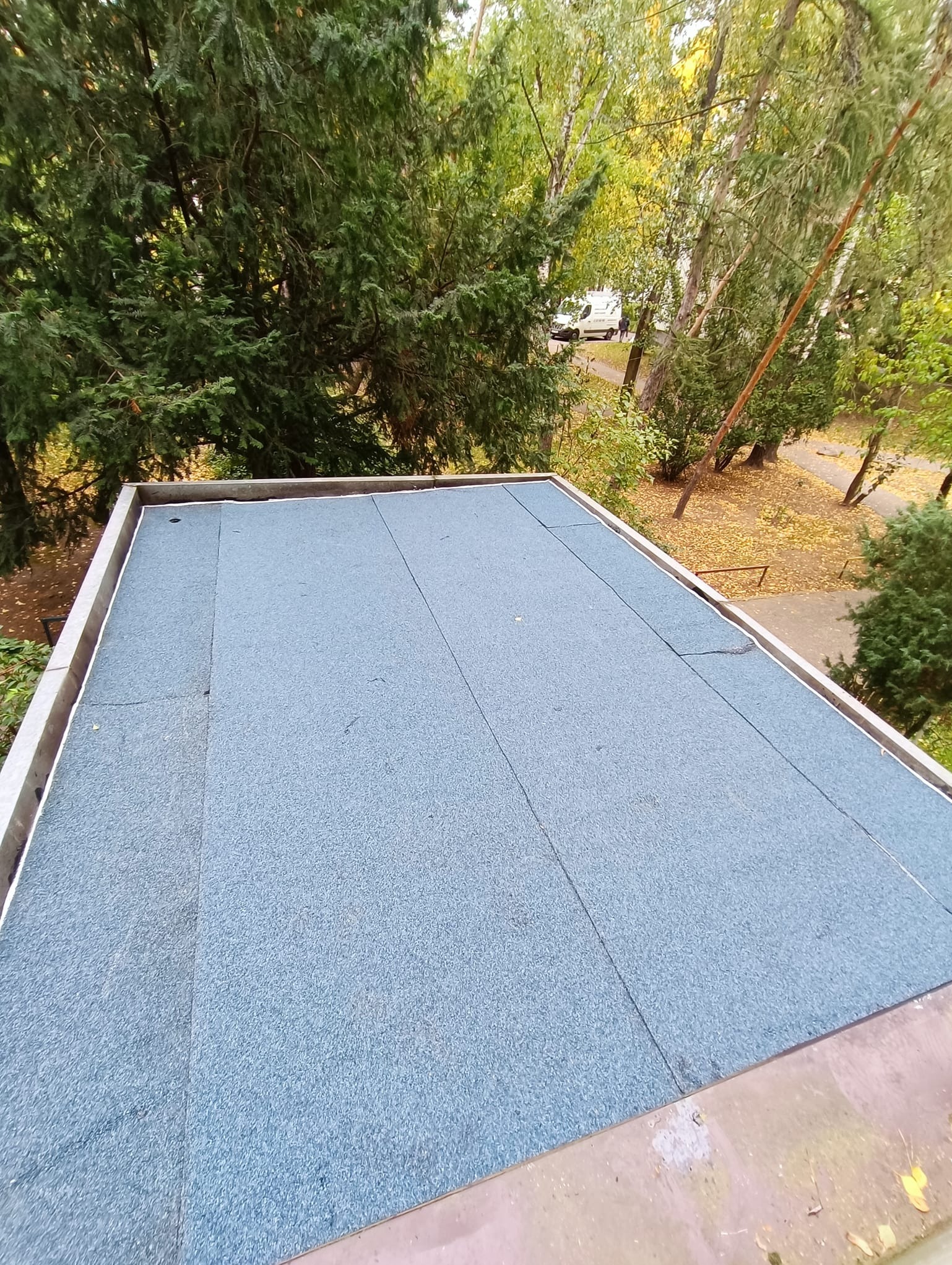 Niebieska, płaska powierzchnia dachu pokryta papą, widok z góry, otoczona zielenią drzew i krzewów w tle.