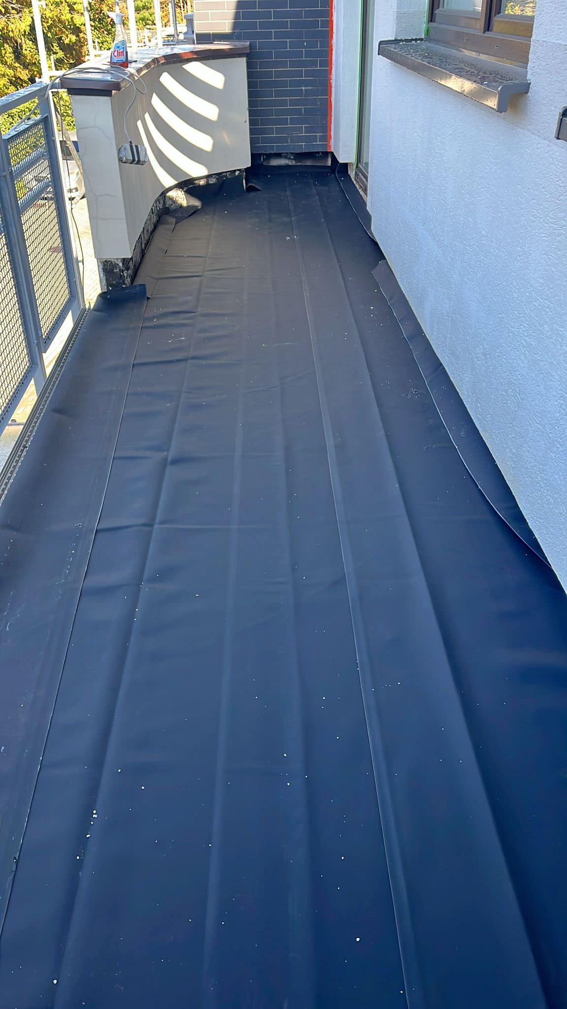 Balkon w trakcie prac zabezpieczających, pokryty czarną folią hydroizolacyjną z widocznymi zakładkami, przygotowany pod dalsze prace wykończeniowe, z widoczną butelką płynu do mycia okien...
