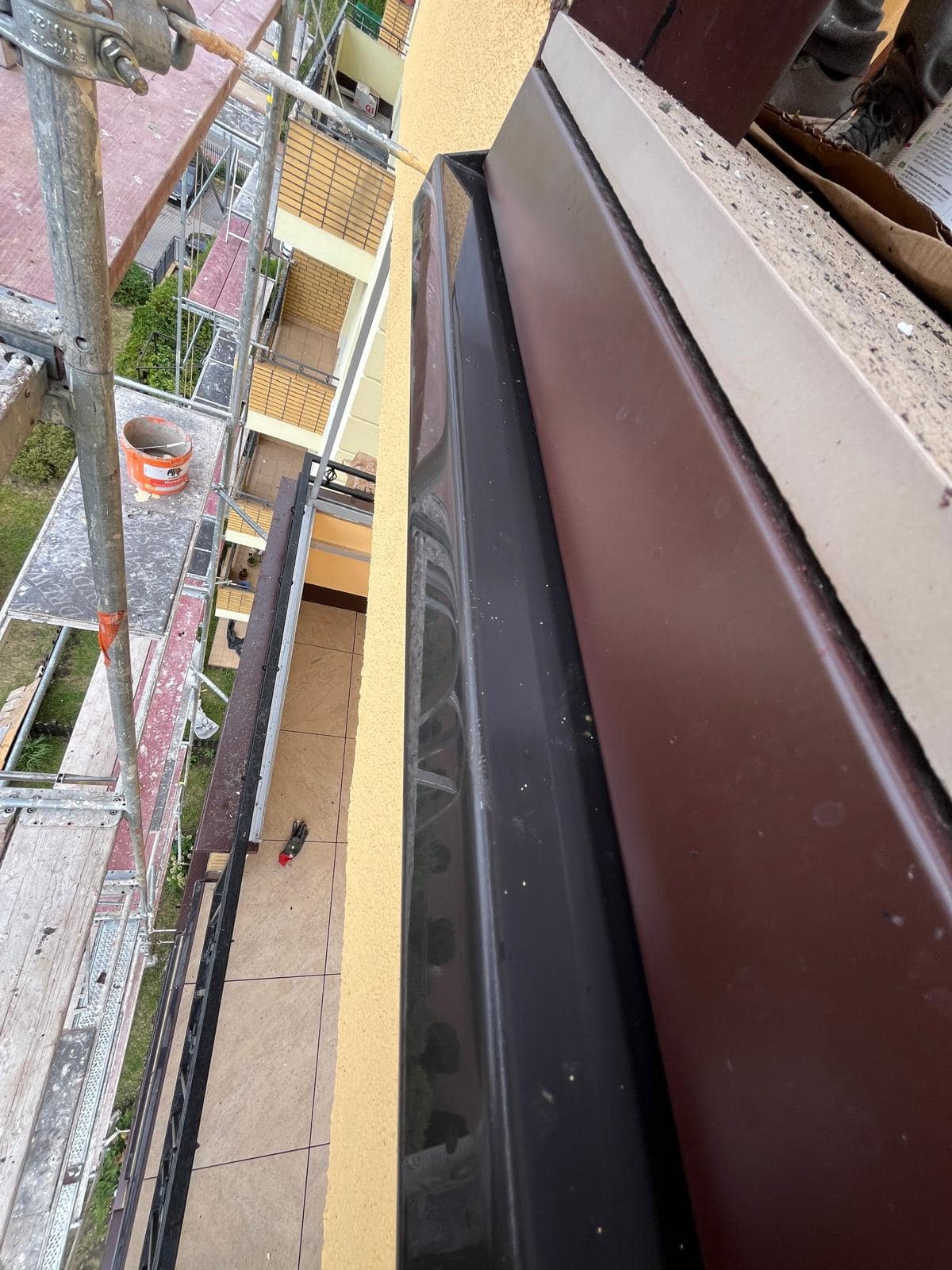 Widok z góry na rusztowanie przy budynku z brązowym wykończeniem elewacji i czarną rynną, z widocznym fragmentem balkonu i narzędziami.