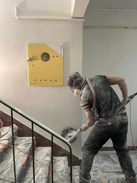 Pracownik w masce szlifuje ścianę na klatce schodowej przy użyciu szlifierki oscylacyjnej, obok skrzynka zabezpieczona przed zabrudzeniem, schody zabezpieczone folią malarską.