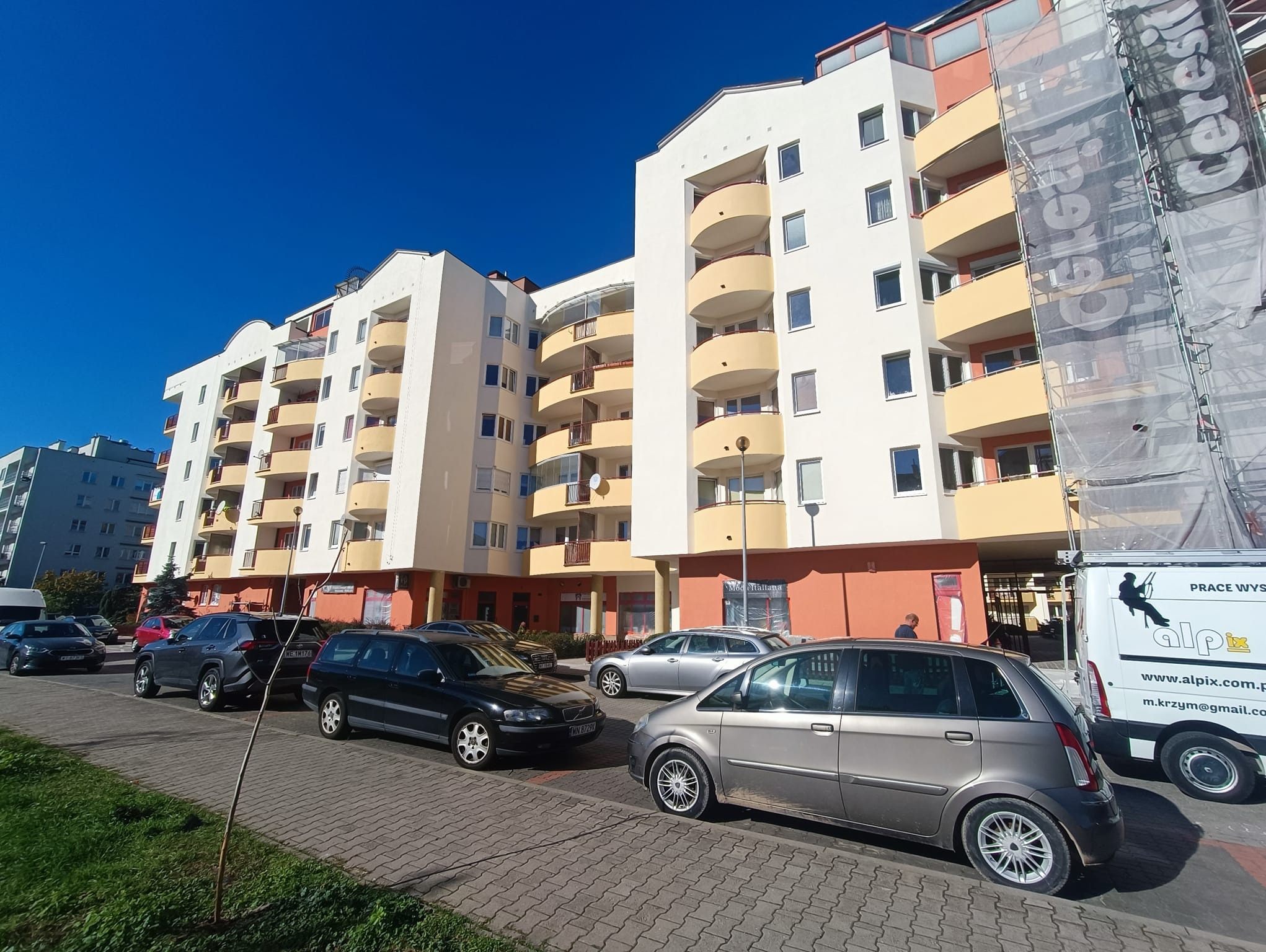 Wielopiętrowy budynek mieszkalny z balkonami w trakcie prac elewacyjnych, widoczne rusztowania z siatką ochronną i zaparkowane samochody na pierwszym planie.
