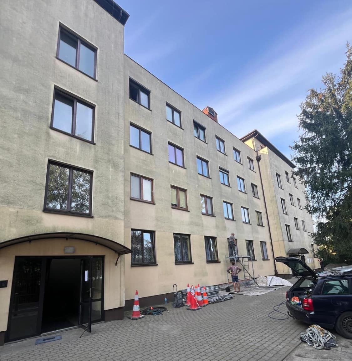 Prace na wysokości przy budynku wielorodzinnym: robotnik na rusztowaniu, widoczne zabezpieczenia, stożki ostrzegawcze i samochód z otwartym bagażnikiem.