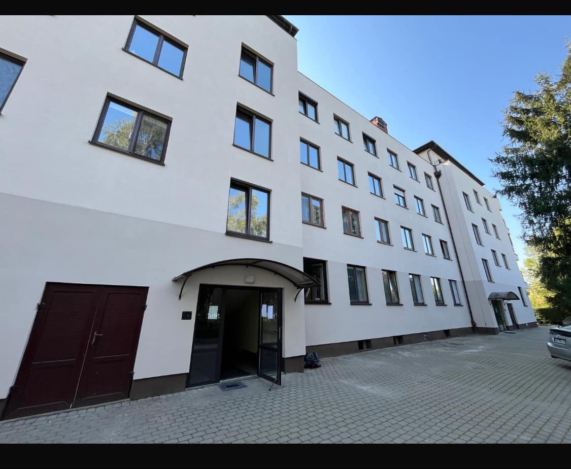 Wielopiętrowy budynek mieszkalny z jasną elewacją, brązowymi ramami okiennymi i wejściem z daszkiem, widoczny z poziomu chodnika z kostki brukowej.