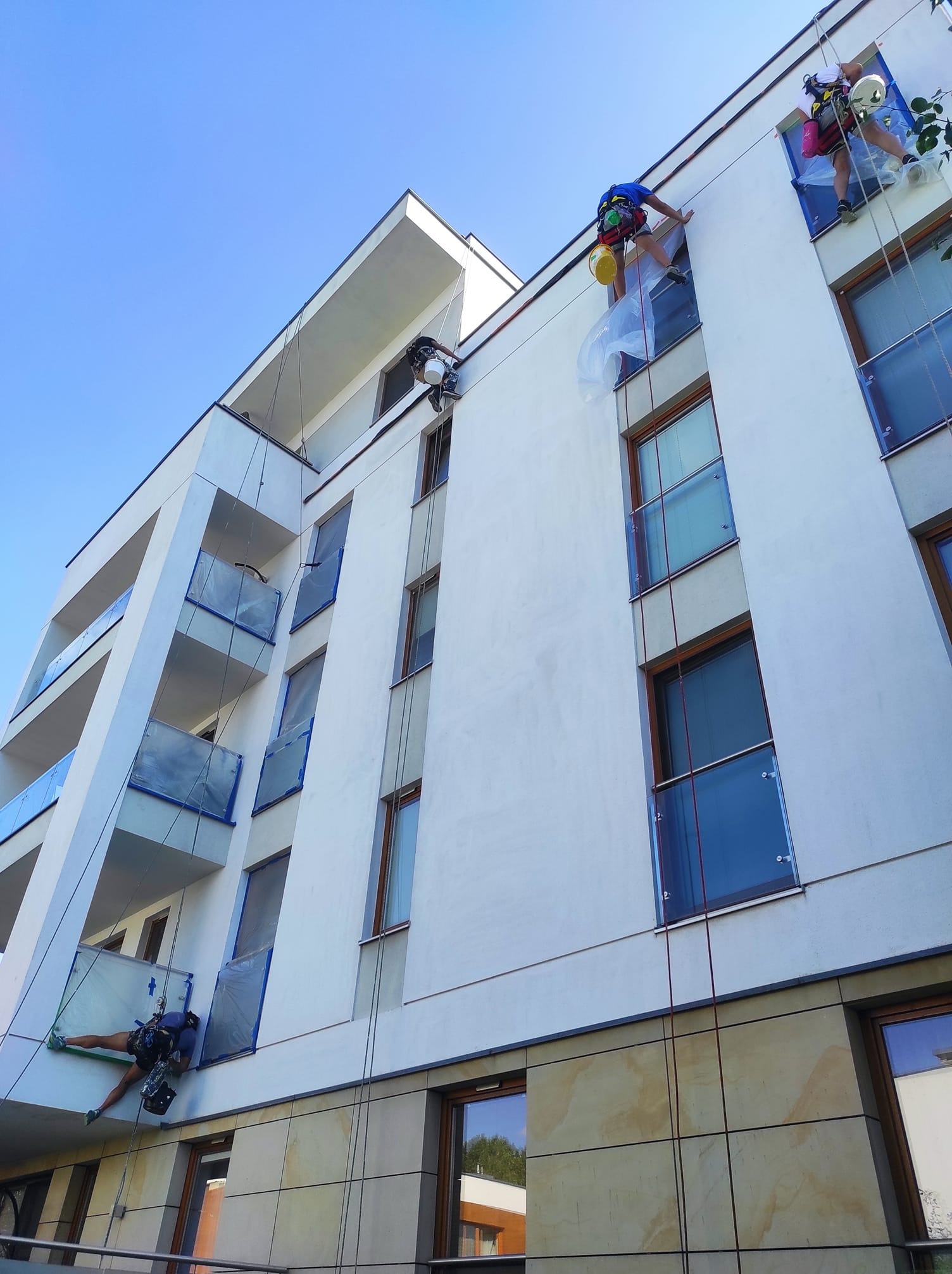 Alpiniści zabezpieczają okna folią przed malowaniem elewacji nowoczesnego budynku z balkonami, widok z dołu na błękitne niebo.