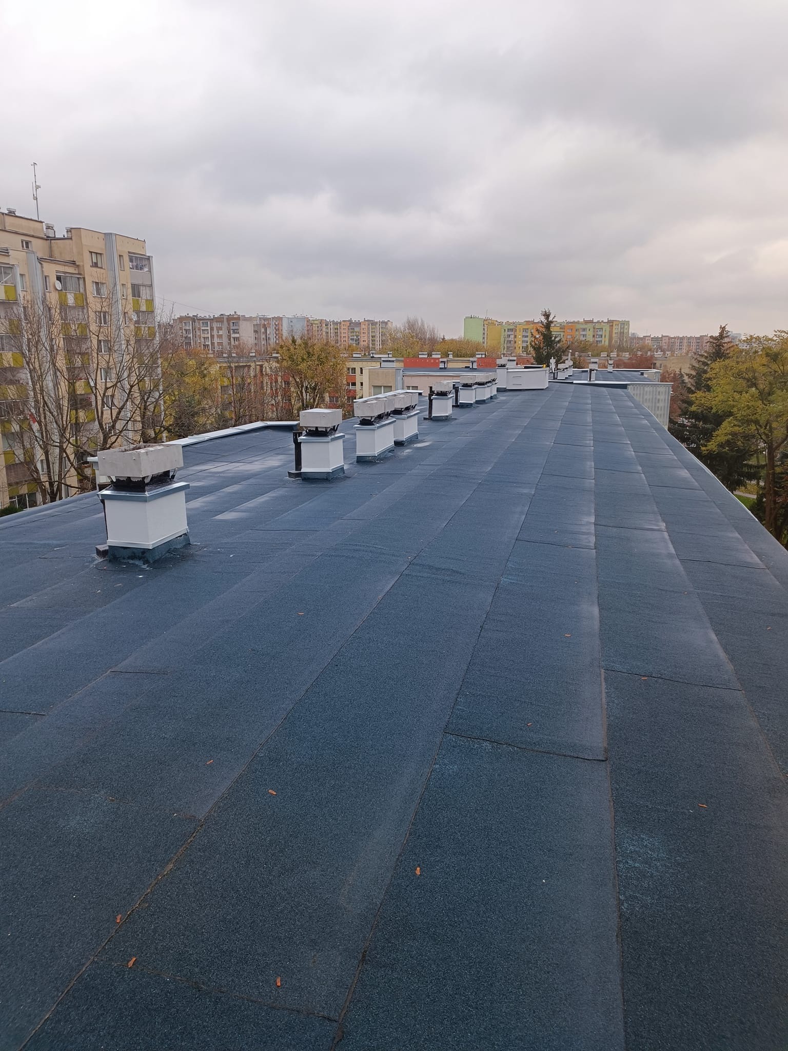Płaski dach pokryty ciemną papą z widocznymi kominkami wentylacyjnymi, w tle bloki mieszkalne i pochmurne niebo.