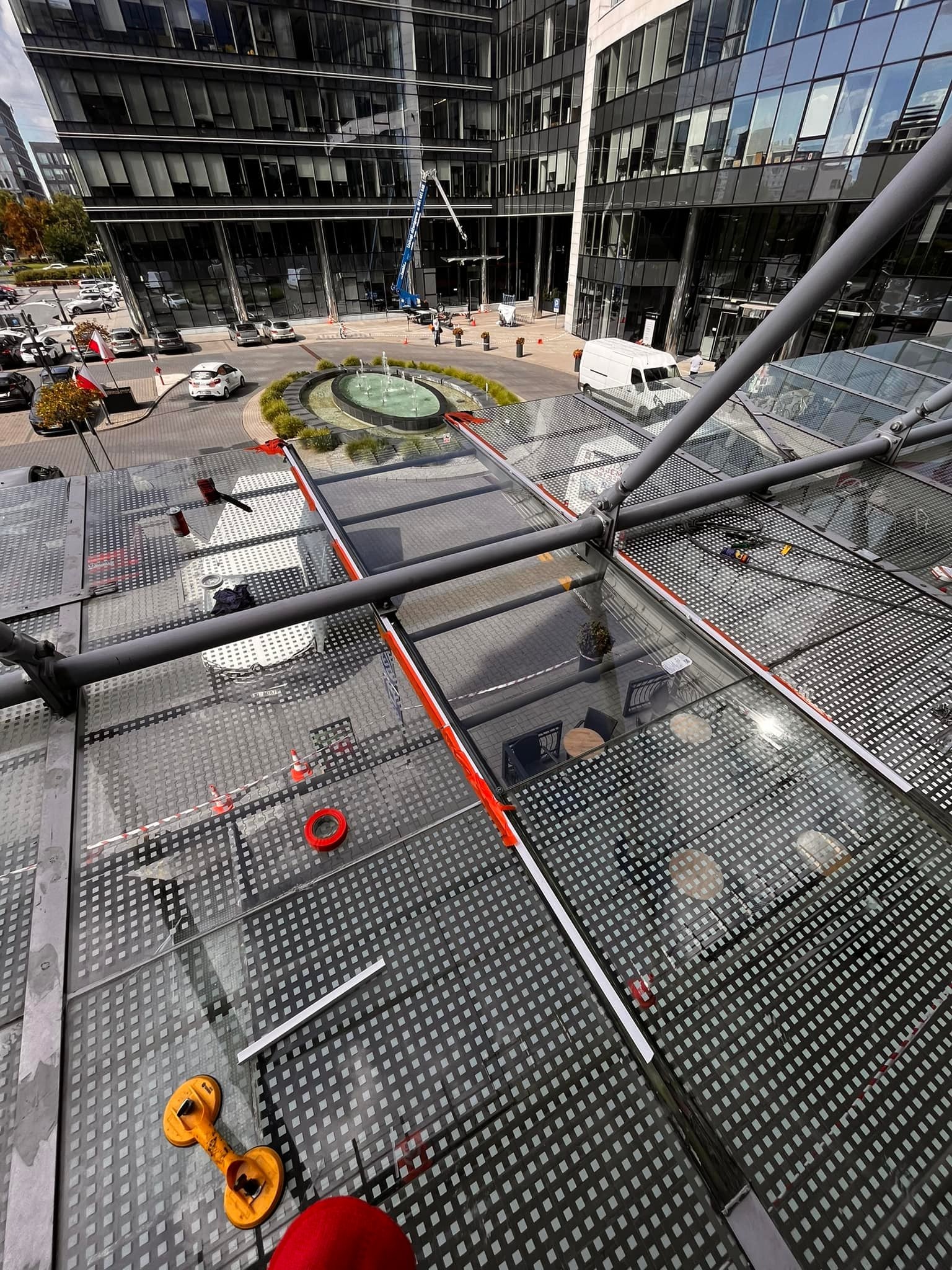 Szklany dach nad pasażem w nowoczesnym kompleksie biurowym, widoczne narzędzia pracy, taśma ostrzegawcza i fontanna w tle.