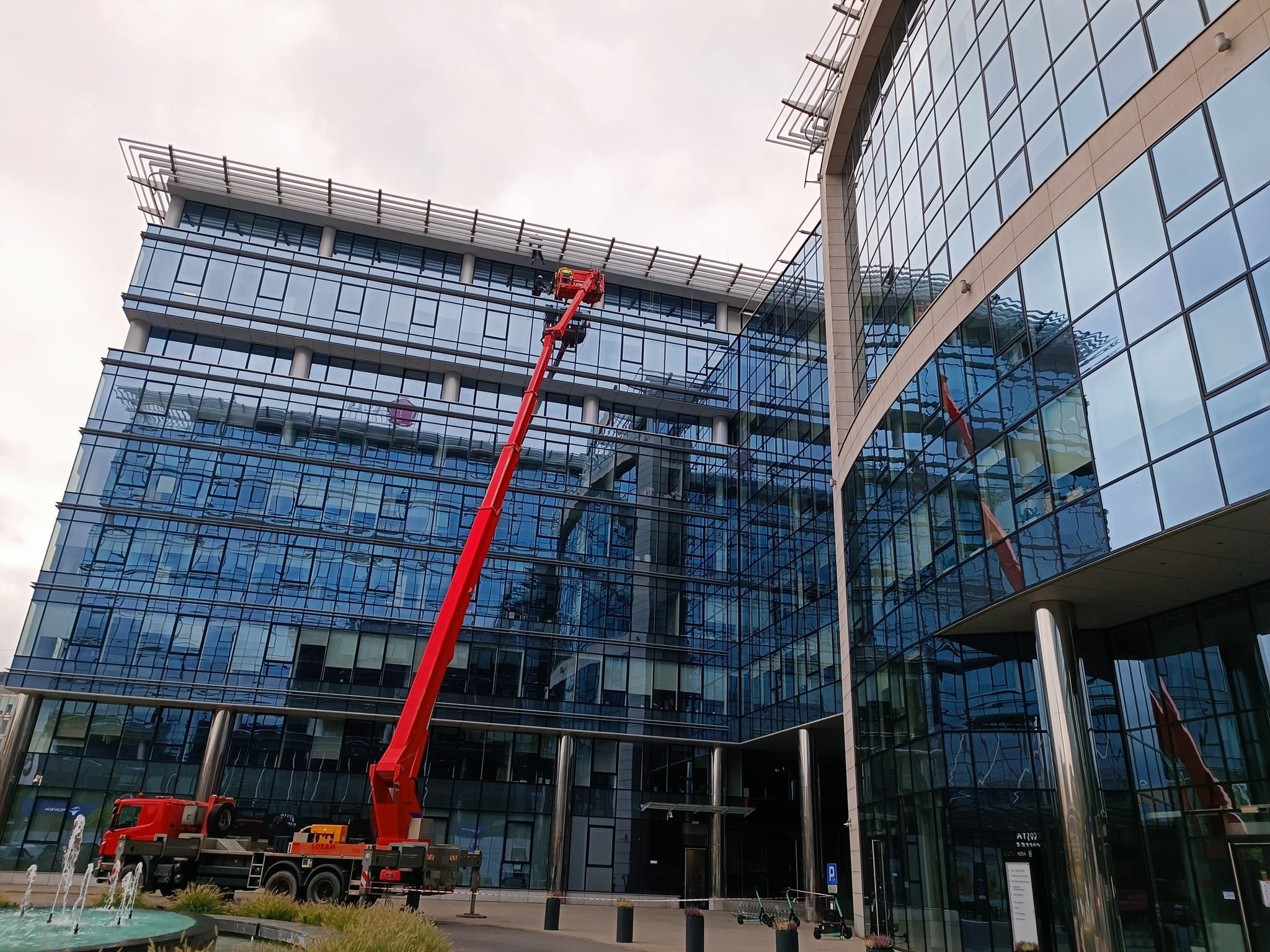 Czerwony podnośnik koszowy na tle nowoczesnego biurowca z szklaną fasadą, dwóch pracowników w koszu na wysokości okien, obok fontanna