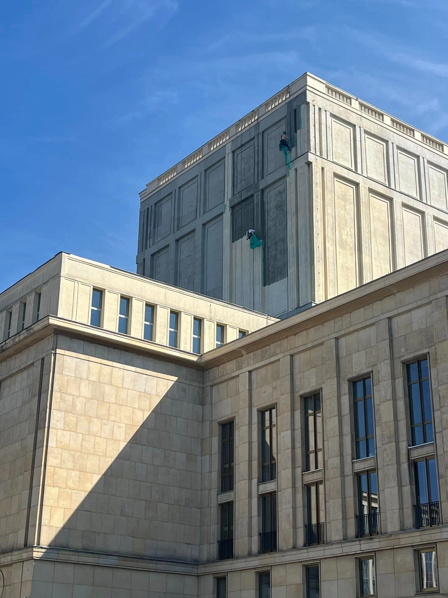 Dwóch alpinistów na linach, zabezpieczonych siatką, pracuje na elewacji wysokiego budynku w stylu modernistycznym. Fragment elewacji jest już odnowiony, ma jaśniejszy odcień. Widoczne są okna...