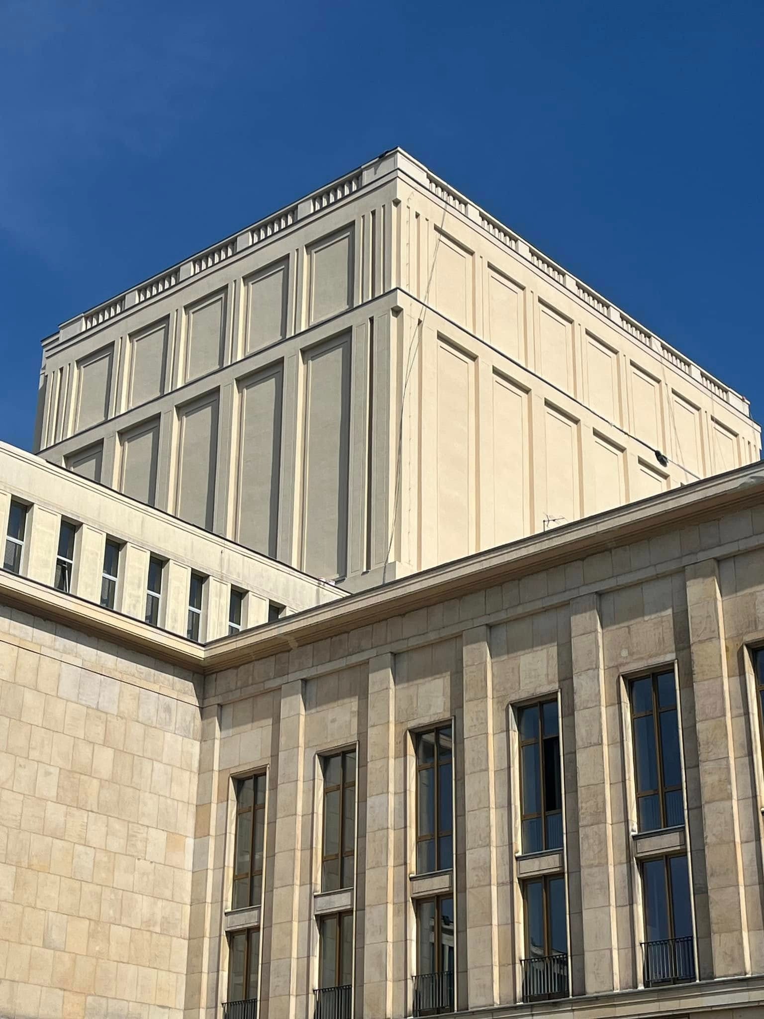 Fragment fasady budynku z widocznymi oknami i dekoracyjnymi elementami architektonicznymi, sfotografowany pod kątem w słoneczny dzień.