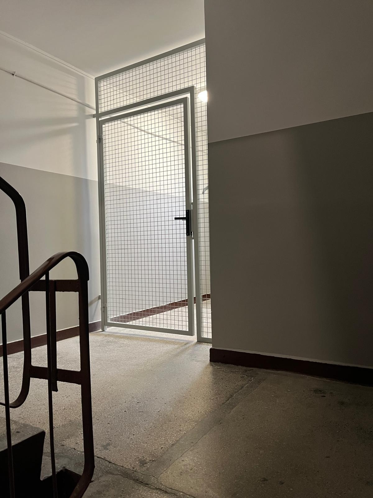 Klatka schodowa z metalową kratą zabezpieczającą wejście, widok z dołu schodów. Szare ściany z ciemniejszym pasem u dołu. Elementy balustrady po lewej stronie.