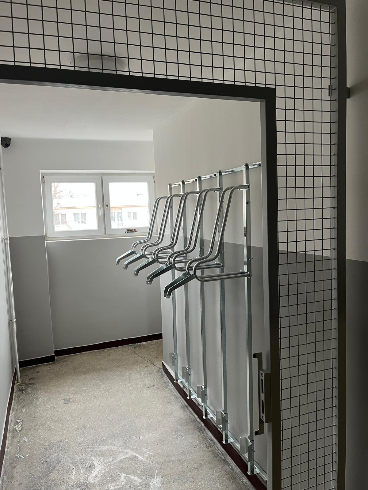 Metalowa konstrukcja z uchwytami na rowery w szarym pomieszczeniu, widoczna przez metalową kratę. Minimalistyczny design, surowe wykończenie, widok z perspektywy wejścia.