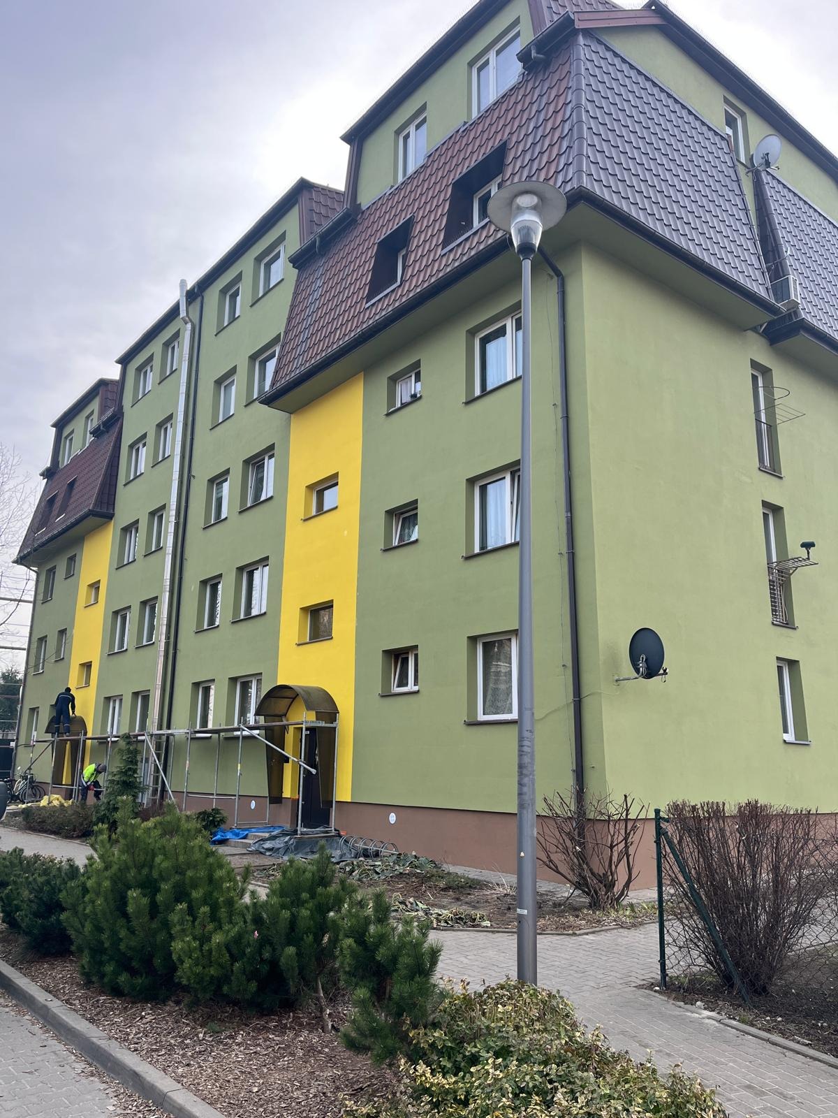 Wielopiętrowy budynek po malowaniu elewacji, z fragmentami w kolorze żółtym i zielonym. Widoczne rusztowania i pracownicy podczas wykonywania prac malarskich.