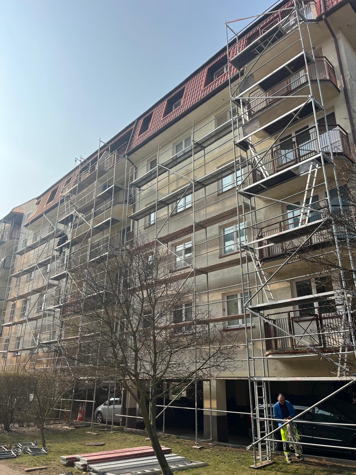 Wielopiętrowy budynek mieszkalny w trakcie prac elewacyjnych, osłonięty rusztowaniami. Widoczny pracownik w odblaskowej odzieży. Słoneczny dzień.
