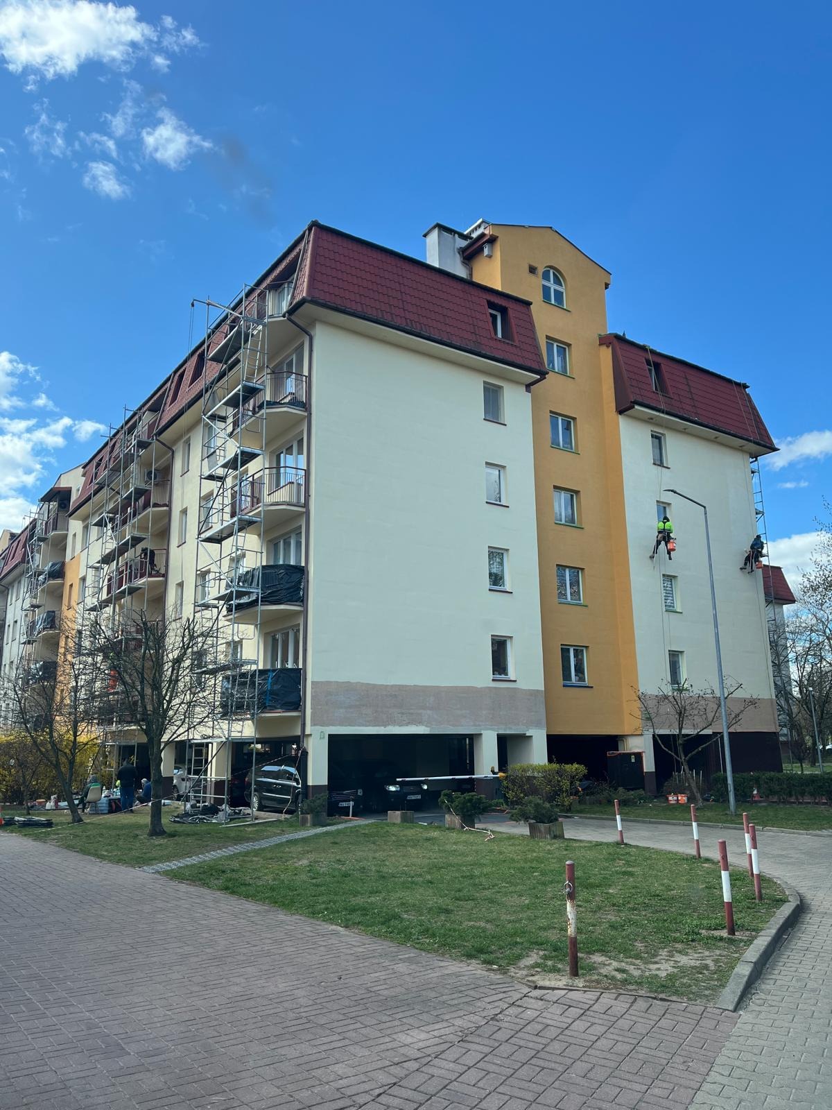 Wielopiętrowy budynek z rusztowaniami i pracownikami na linach podczas malowania elewacji. Widoczne fragmenty pomalowanej ściany i przygotowane podłoże.