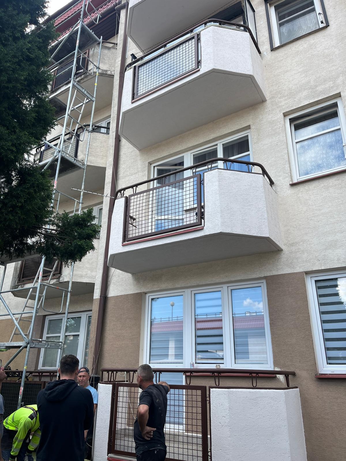 Budynek z balkonami w trakcie prac. Widoczne rusztowania i ekipa remontowa. Jasna elewacja z brązowym pasem. Metalowe balustrady balkonowe.