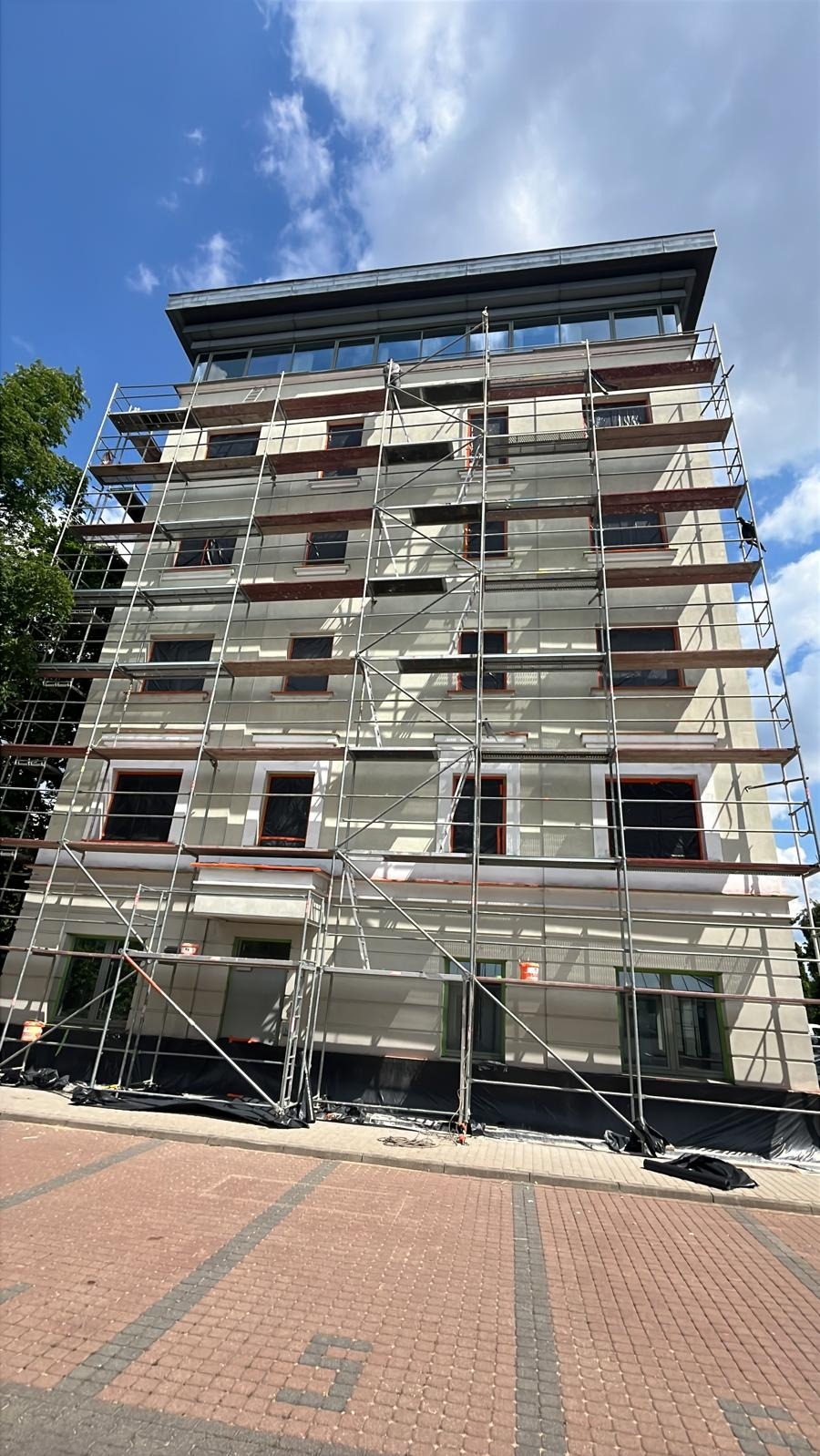 Wielopiętrowy budynek w trakcie prac elewacyjnych, osłonięte okna, rusztowania na całej wysokości, widok z dołu, błękitne niebo z chmurami w tle.