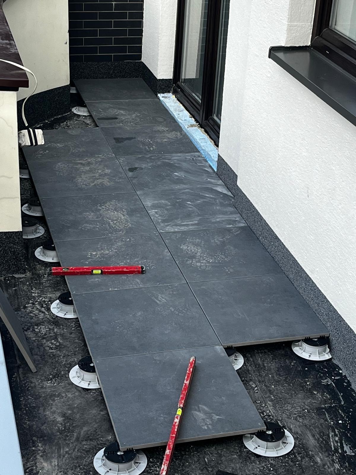 Układanie ciemnoszarych płytek na regulowanych wspornikach na balkonie. Poziomica na płytkach. Widoczne zabezpieczenie przy oknie balkonowym.