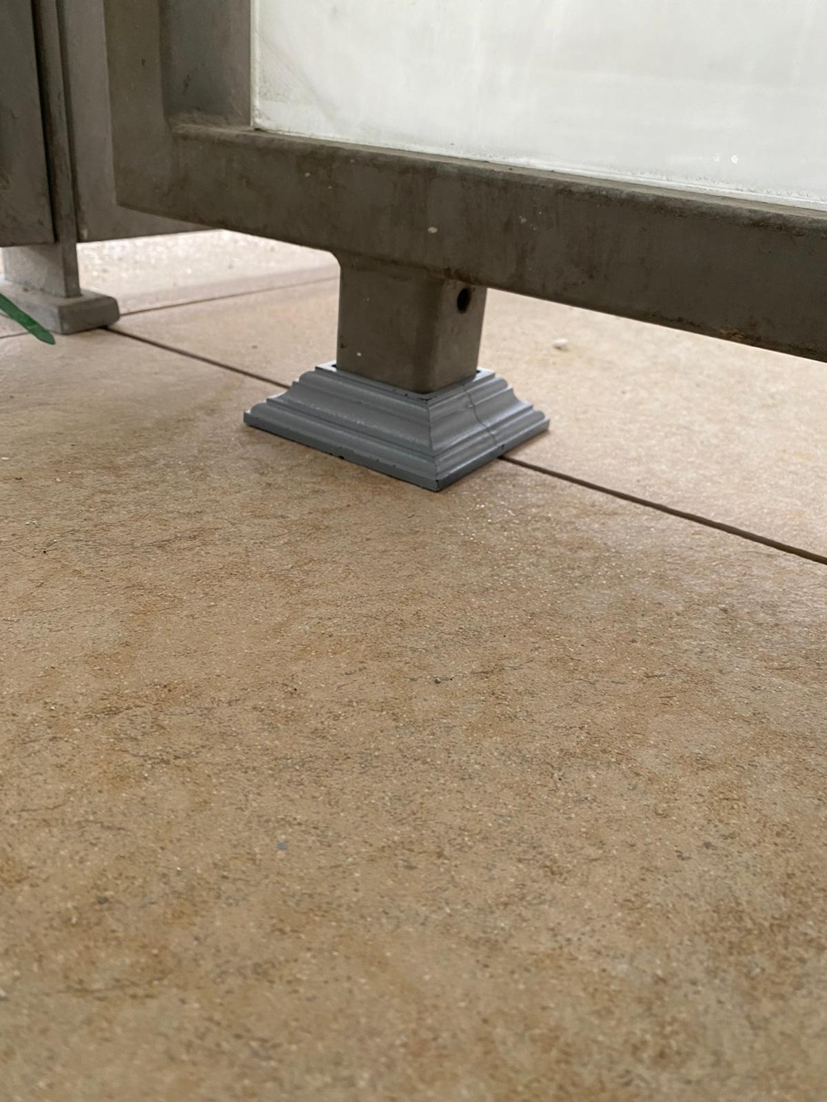 Szary, pomalowany wspornik balustrady balkonowej z widocznymi śladami użytkowania, osadzony na brązowej, ceramicznej posadzce balkonu. Ujęcie z dołu.