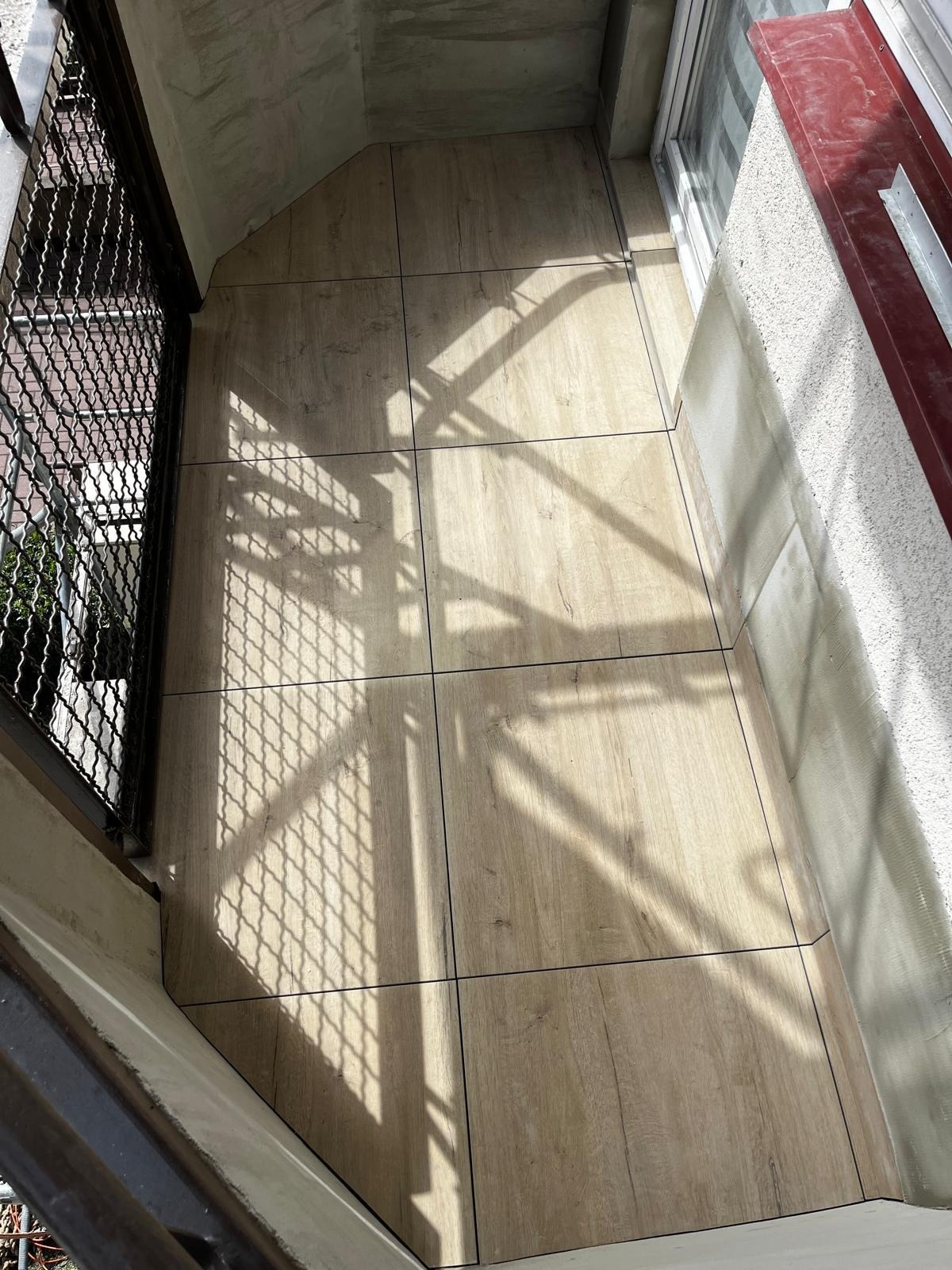 Wykończony balkon z nową, jasną podłogą z płytek imitujących drewno, widok z góry, cień kraty balkonowej. Estetyczne połączenie z elewacją.