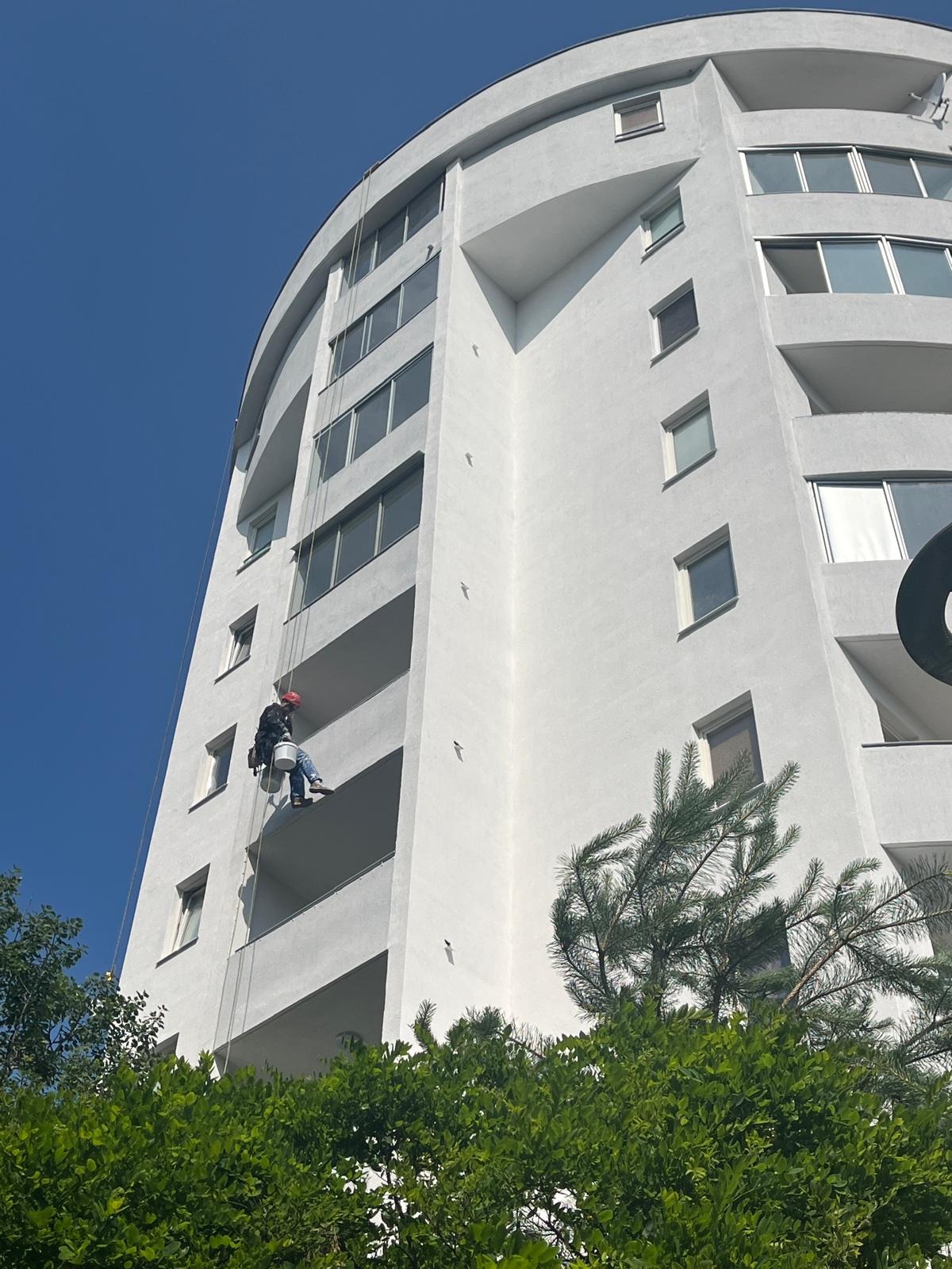 Pracownik na linach, w kasku, maluje białą elewację wysokiego budynku z balkonami. Widok z dołu, błękitne niebo w tle, zieleń drzew w dolnej części kadru.
