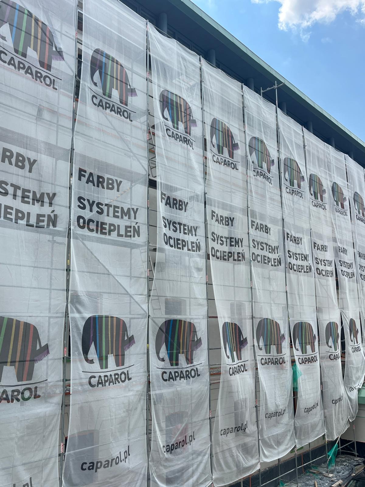 Elewacja budynku w trakcie ocieplania, osłonięta siatką z logo Caparol. Widoczne rusztowania i fragment nieba. Prace ociepleniowe w toku.