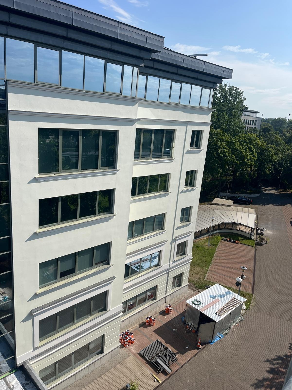 Jasna elewacja budynku biurowego z widocznymi oknami, rusztowaniem i pojemnikami z farbą. Ujęcie z góry, w tle zieleń drzew i fragment innego budynku.