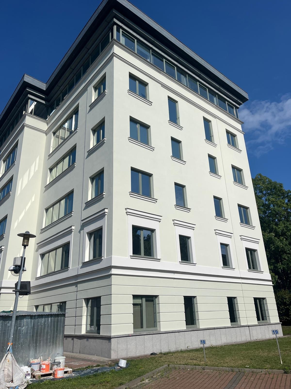 Odnowiona elewacja budynku biurowego w jasnych barwach, z widocznymi detalami architektonicznymi i świeżo pomalowanymi oknami. Na dole widoczne farby i numeracja.