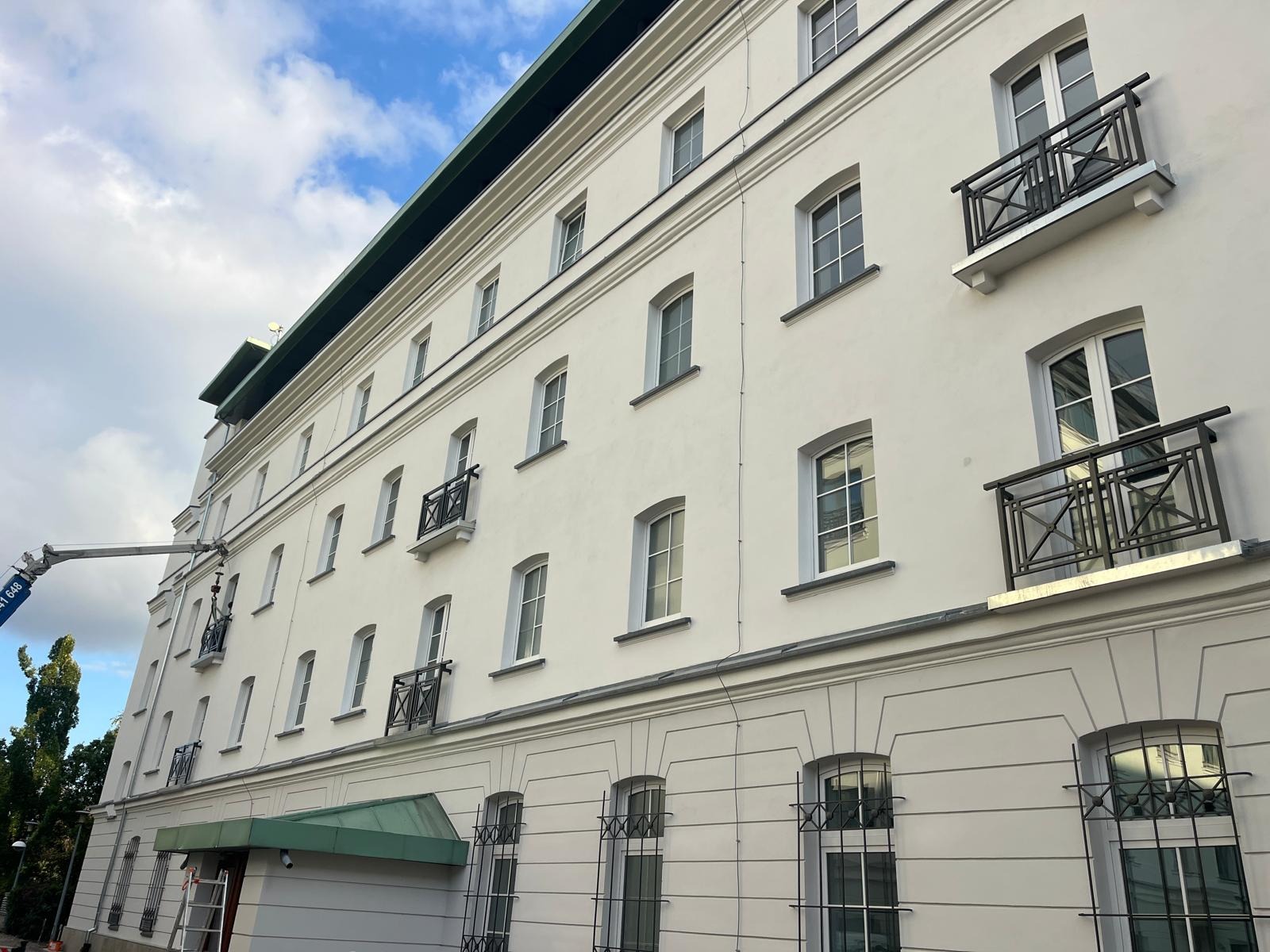 Biała elewacja budynku po renowacji w Starych Babicach, widoczna z podnośnika koszowego. Balkony z metalowymi balustradami i okna z kratami.