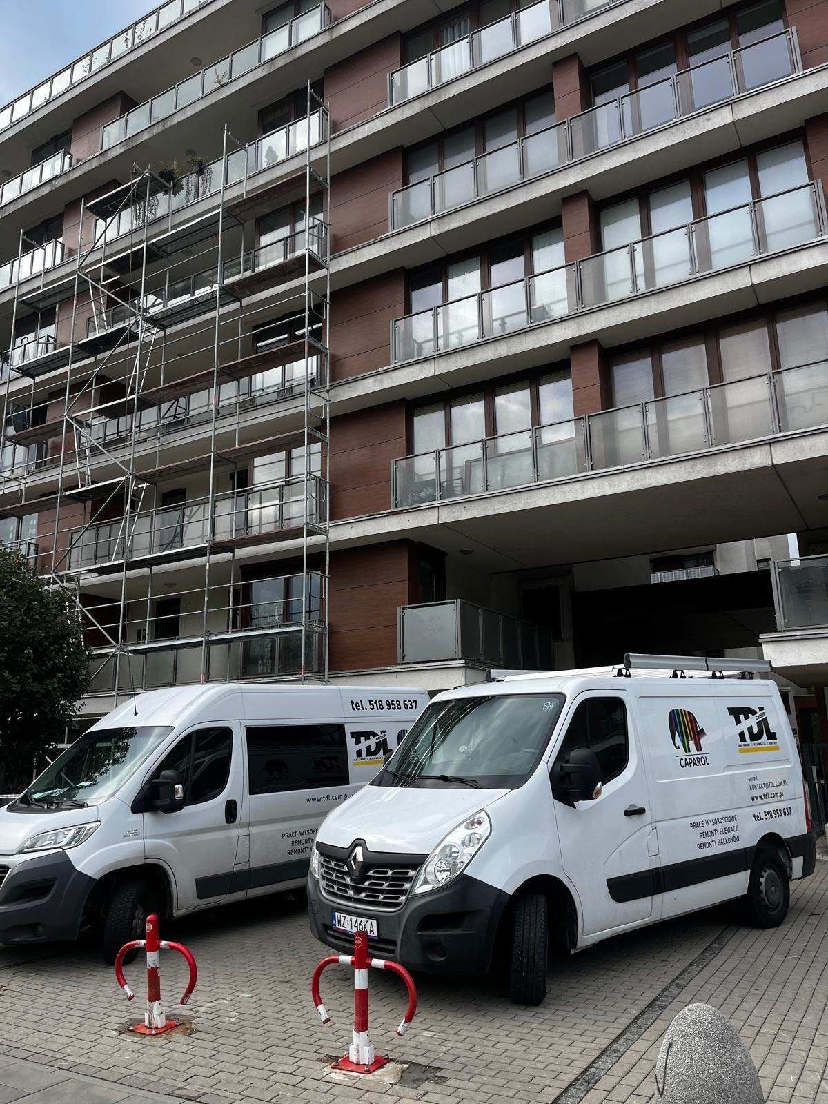 Wielopiętrowy budynek z balkonami w trakcie prac elewacyjnych, widoczne rusztowania i dwa białe busy z logo firmy wykonawczej na pierwszym planie.