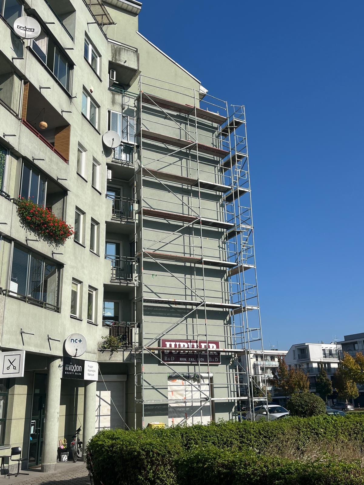 Budynek w rusztowaniach, widoczne anteny satelitarne i balkony z kwiatami. Część elewacji zakryta siatką. Błękitne niebo w tle. Ujęcie z poziomu ulicy.