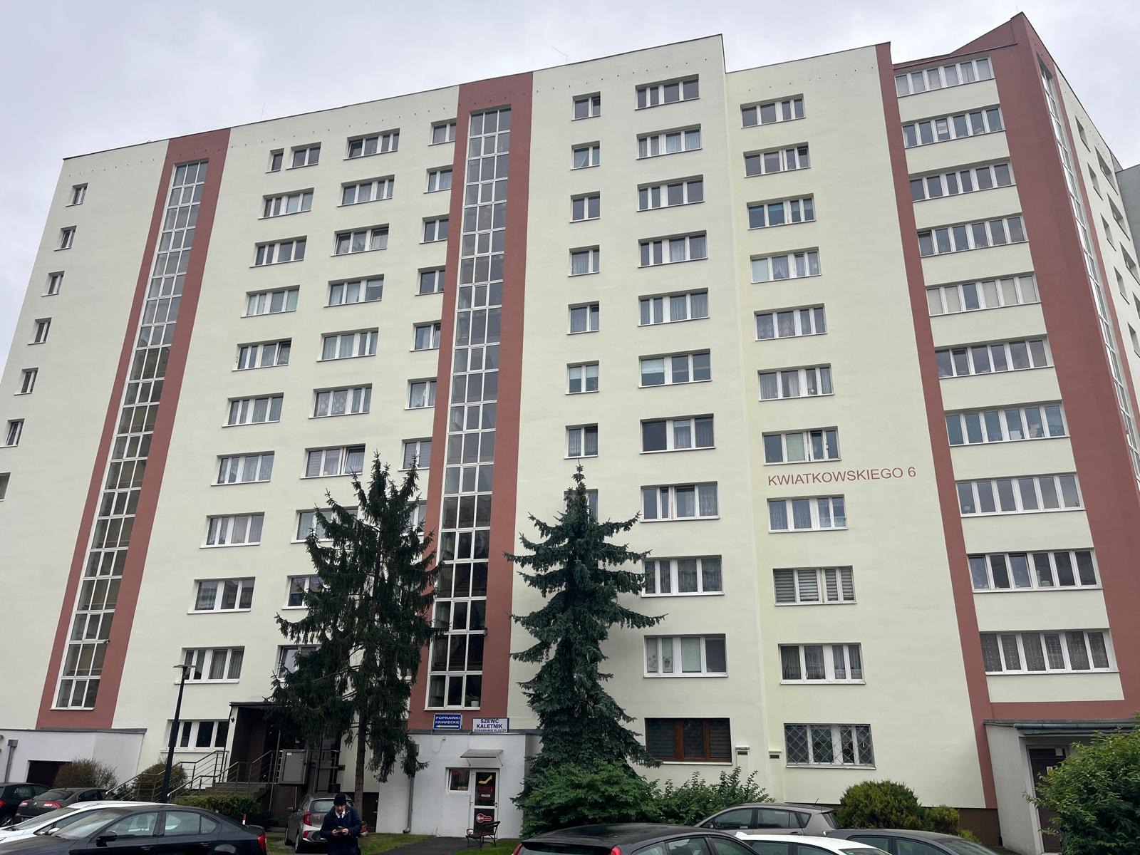 Wysoki budynek mieszkalny z jasną elewacją i czerwonymi akcentami, widoczne liczne okna i wejście. Dwa wysokie drzewa iglaste przed budynkiem. Tabliczka z adresem.