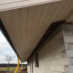 Drewniana podbitka dachowa z widocznymi łączeniami desek, czarna rynna i fragment ściany z białych bloczków betonowych w surowym stanie, na tle pochmurnego nieba i krajobrazu.