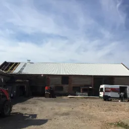 Budynek gospodarczy z częściowo zdjętym i nowym, metalowym dachem, widoczny wóz serwisowy z logo firmy dekarskiej i niebieskie niebo w tle.