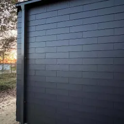 Fragment elewacji budynku pokrytej ciemnoszarymi panelami imitującymi łupek, z widoczną rynną spustową i drzewem w tle o zachodzie słońca.