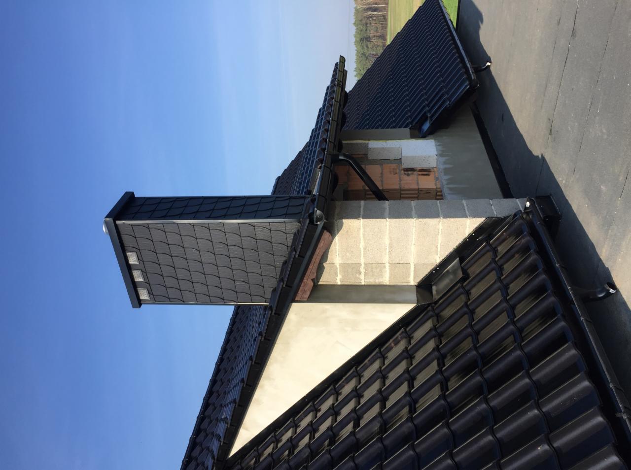 Fragment dachu z czarną dachówką ceramiczną, widoczny komin z szarej dachówki, orynnowanie i fragment ściany w trakcie prac budowlanych, na tle błękitnego nieba.