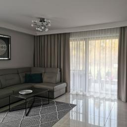 Fom-artdeconstruire-Adam Blachewicz - Jasne wnętrze salonu z szarym narożnikiem, dywanem w romby, błyszczącymi płytkami podłogowymi i zasłonami w odcieniach szarości oraz białymi firanami, widok z okna na zewnątrz.