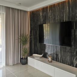 Fom-artdeconstruire-Adam Blachewicz - Salon z marmurową ścianą, na której wisi telewizor, biała szafka RTV z dekoracjami, roślina w doniczce i szare zasłony przy oknie.