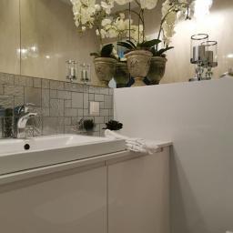 Fom-artdeconstruire-Adam Blachewicz - Nowoczesna łazienka z białą umywalką nablatową, srebrną mozaiką na ścianie, białymi frontami szafek i dekoracjami z kwiatów orchidei w ozdobnych doniczkach.