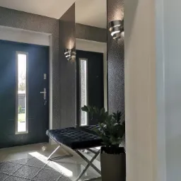 Nowoczesny przedpokój z szarą tapetą w geometryczny wzór, lustrem, czarną ławką z chromowanymi nogami, dopasowanym dywanem i rośliną w czarnej donicy, oświetlony spiralnymi kinkietami.