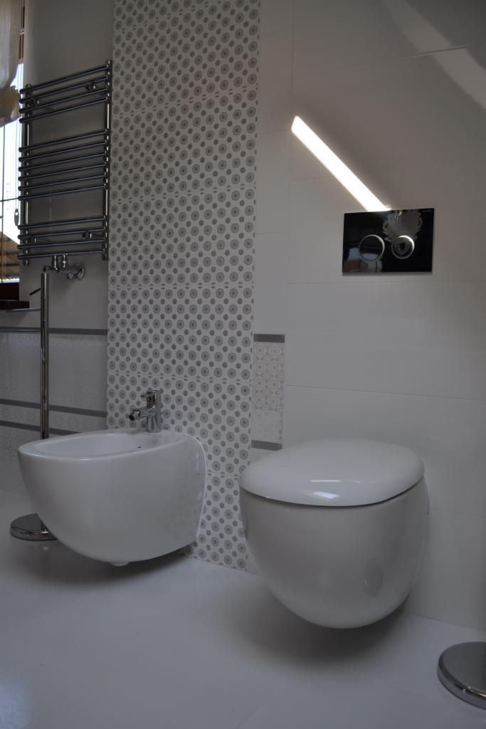 Nowoczesna łazienka w bieli: podwieszana toaleta i bidet obok grzejnika drabinkowego na tle ściany z geometrycznym wzorem. Minimalistyczny design i czystość.