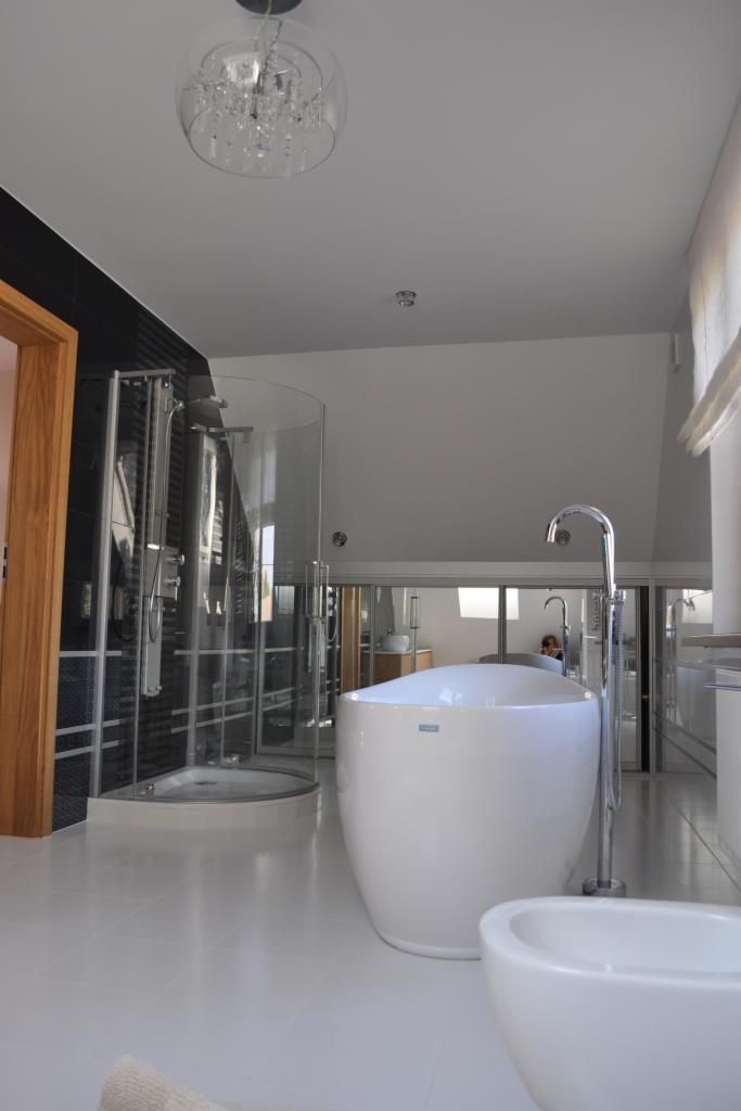 Jasna łazienka z kabiną prysznicową typu walk-in, wolnostojącą wanną i bidetem. Na suficie widoczny ozdobny żyrandol. Minimalistyczny wystrój, białe płytki podłogowe.