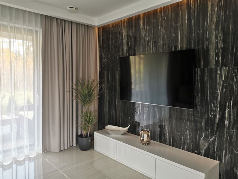 Salon z marmurową ścianą, na której wisi telewizor, biała szafka RTV z dekoracjami, roślina w doniczce i szare zasłony przy oknie.