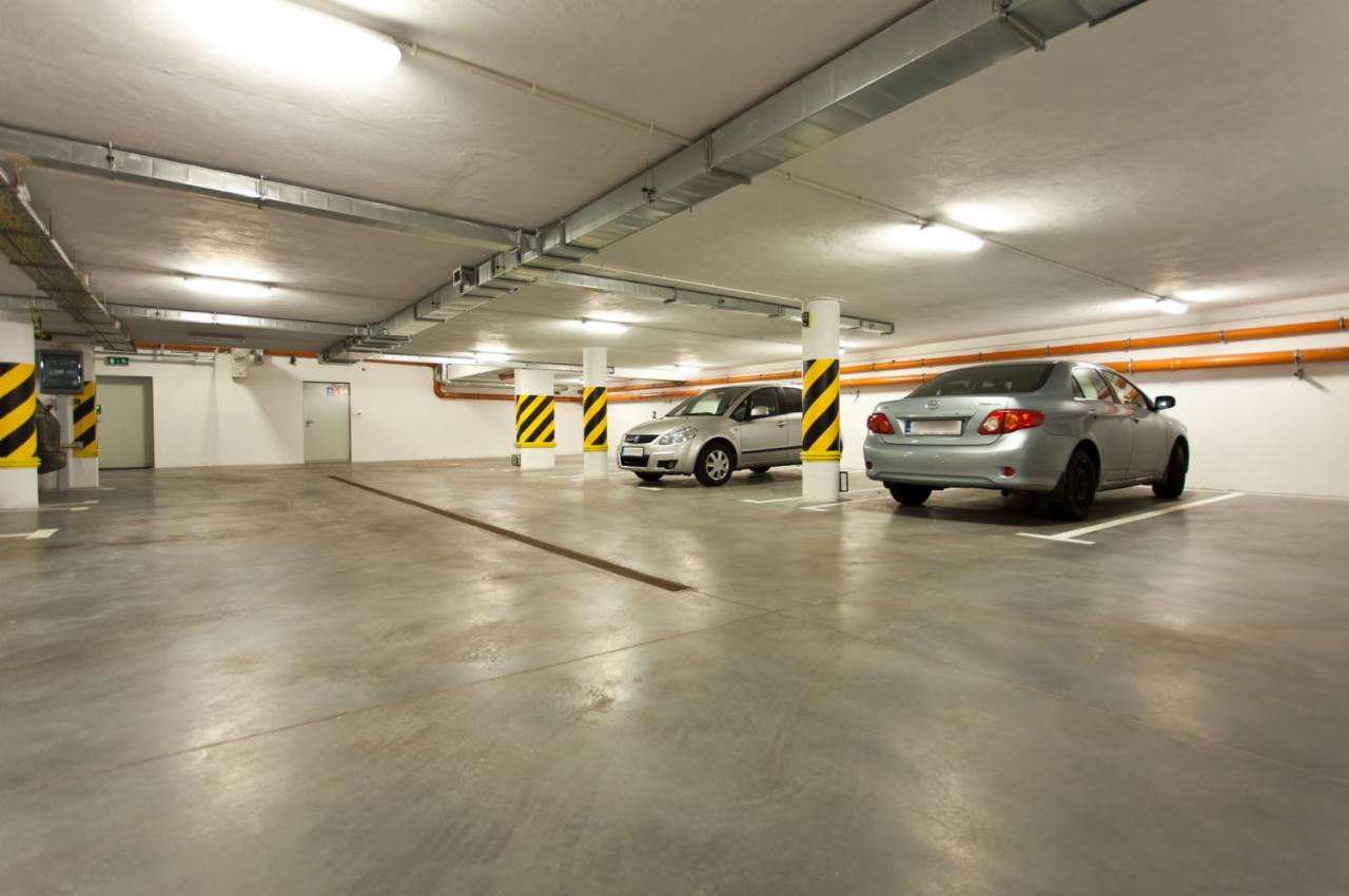 Wnętrze parkingu podziemnego z dwoma zaparkowanymi, srebrnymi samochodami. Kolumny oznaczone pasami ostrzegawczymi. Dobre oświetlenie.