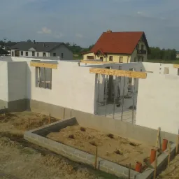 Surowy stan domu z białych bloczków, widoczne fundamenty i ściany parteru. W tle zabudowa jednorodzinna i błękitne niebo.