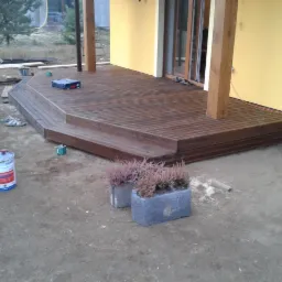 Drewniany taras przy żółtej ścianie domu, z widocznymi narzędziami i farbą. Ukończona konstrukcja z desek, z dwupoziomowym podestem i elementami roślinnymi w donicach.