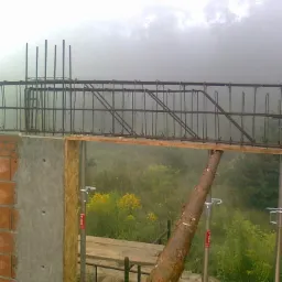 Zbrojenie nadproża z ceglaną ścianą i podporami na tle mglistego krajobrazu. Widoczne żółte kwiaty i fragment drewnianego szalunku.