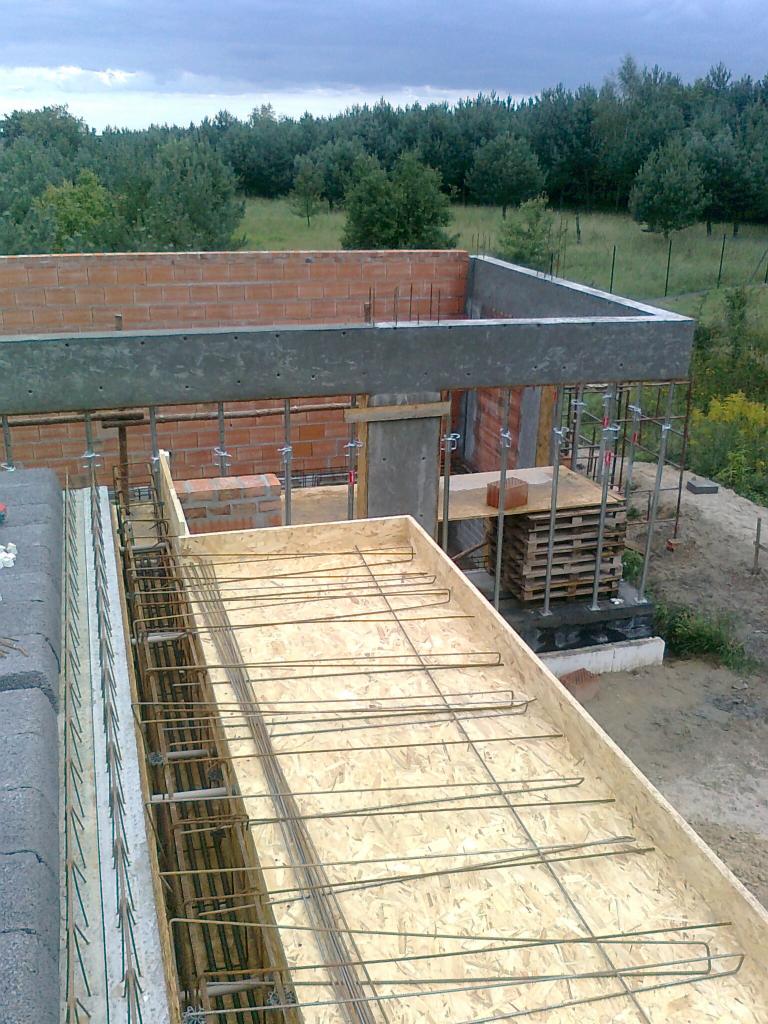 Budowa domu: zbrojenie stropu w szalunku z desek, widoczny mur z cegły i betonowy wieniec. W tle drzewa i pochmurne niebo. Etap prac budowlanych.